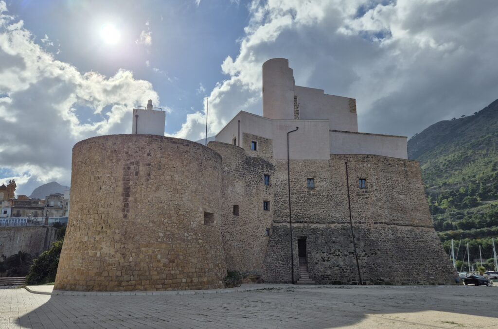 Castellammare Castle Exterior: The majestic Arab-Norman castle of Castellammare del Golfo, standing proudly by the sea, with its stone walls bathed in golden sunlight and waves gently lapping at its base.